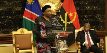 Woman in a patterned dress speaks at a clear podium with microphone, flanked by Namibian flags and a seated man in a suit behind her in a formal government setting