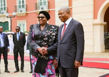Two formally dressed leaders shake hands and smile on a red carpet outside a pastel building.
