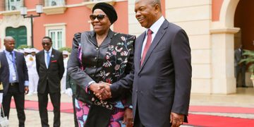 Two formally dressed leaders shake hands and smile on a red carpet outside a pastel building.