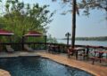 Outdoor pool deck by a river with lounge chairs, tables, and red umbrellas; trees and water in the background.