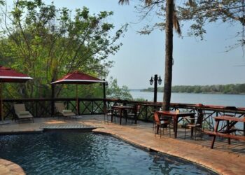 Outdoor pool deck by a river with lounge chairs, tables, and red umbrellas; trees and water in the background.