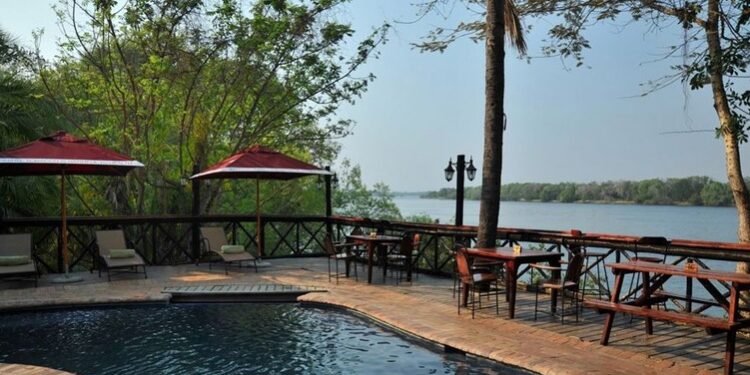 Outdoor pool deck by a river with lounge chairs, tables, and red umbrellas; trees and water in the background.