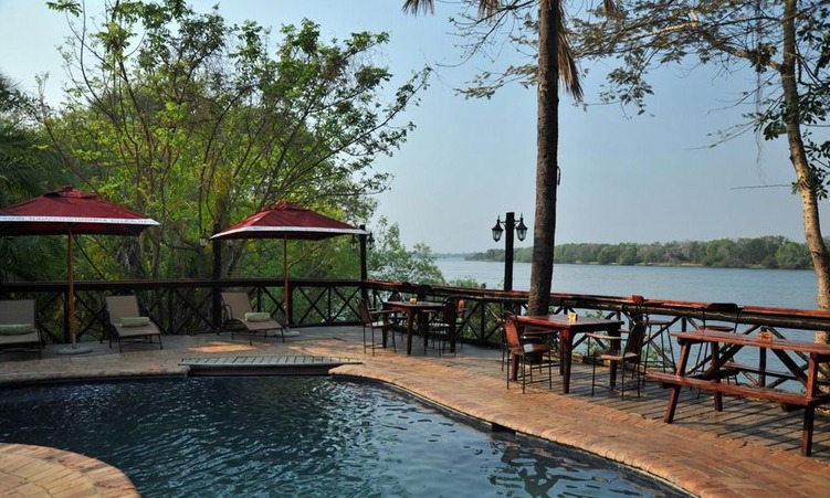 Poolside terrace with red umbrellas, lounge chairs, and a brick deck overlooking a calm river.