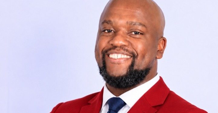 Professional headshot of a smiling man in a red blazer and navy tie against a light background.