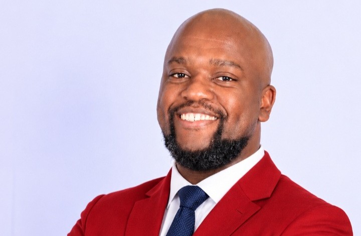 Professional headshot of a smiling man in a red blazer and navy tie against a light background.
