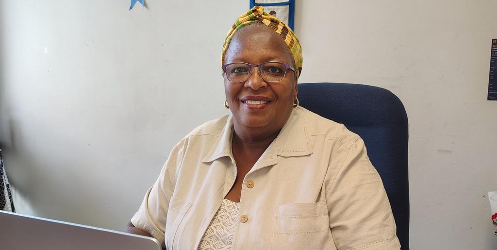 Dr Wanja Njuguna - The Brief | Namibia's Leading Business & Financial News Smiling woman wearing glasses and a colorful head wrap sits at a desk with a laptop in a beige shirt, in an office setting.