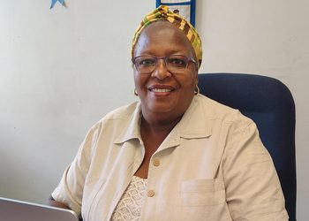 Smiling woman wearing glasses and a colorful head wrap sits at a desk with a laptop in a beige shirt, in an office setting.