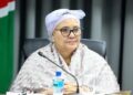 LUCIA WITBOOI - The Brief | Namibia's Leading Business & Financial News Woman wearing a light patterned hijab and glasses sits at a table with a microphone, water bottle, and glass in front, in a conference setting.