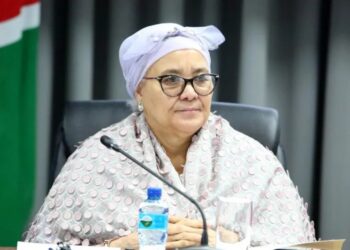LUCIA WITBOOI - The Brief | Namibia's Leading Business & Financial News Woman wearing a light patterned hijab and glasses sits at a table with a microphone, water bottle, and glass in front, in a conference setting.