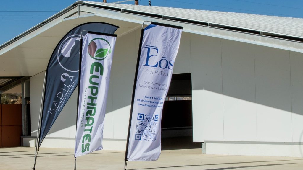 Three vertical banners outside a modern white building; left banner is dark with a logo, center banner has green vertical text, right banner reads 'EOS CAPITAL' with a QR code.