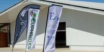 Three vertical banners outside a modern white building; left banner is dark with a logo, center banner has green vertical text, right banner reads 'EOS CAPITAL' with a QR code.