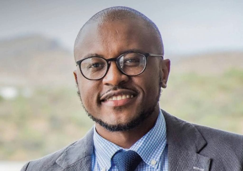 HAFENI KASHIMBONDE - The Brief | Namibia's Leading Business & Financial News Professional headshot of a smiling man wearing round glasses, a gray suit, and a blue check shirt.