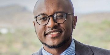 Professional headshot of a smiling man wearing round glasses, a gray suit, and a blue check shirt.