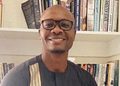 Smiling man wearing glasses stands in front of a bookshelf filled with colorful books.