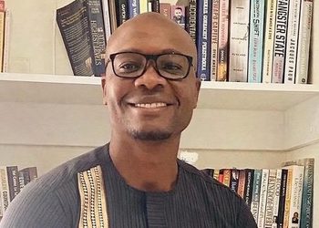 David Ipinge - The Brief | Namibia's Leading Business & Financial News Smiling man wearing glasses stands in front of a bookshelf filled with colorful books.