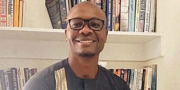Smiling man wearing glasses stands in front of a bookshelf filled with colorful books.