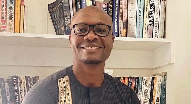 Smiling man wearing glasses stands in front of a bookshelf filled with colorful books.