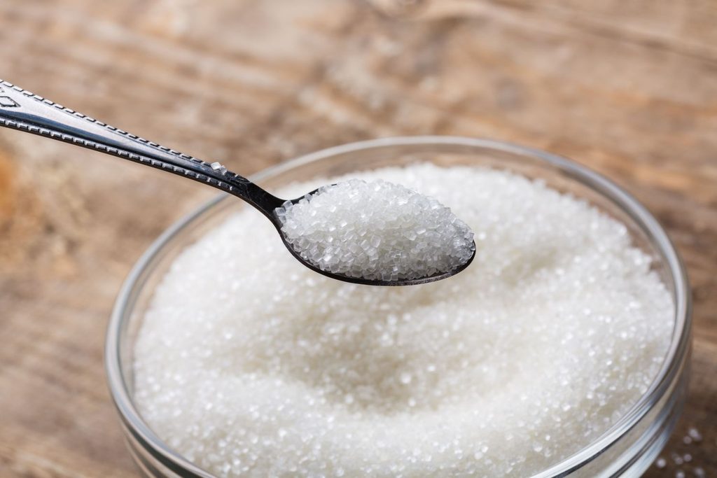 Spoonful of white granulated sugar held over a jar of sugar on a wooden surface