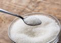 Spoonful of white granulated sugar held over a jar of sugar on a wooden surface