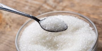 Spoonful of white granulated sugar held over a jar of sugar on a wooden surface