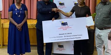 Award ceremony scene: two men shake hands as a woman presents a large check from the Livestock and Livestock Products Board of Namibia, with a banner behind them.