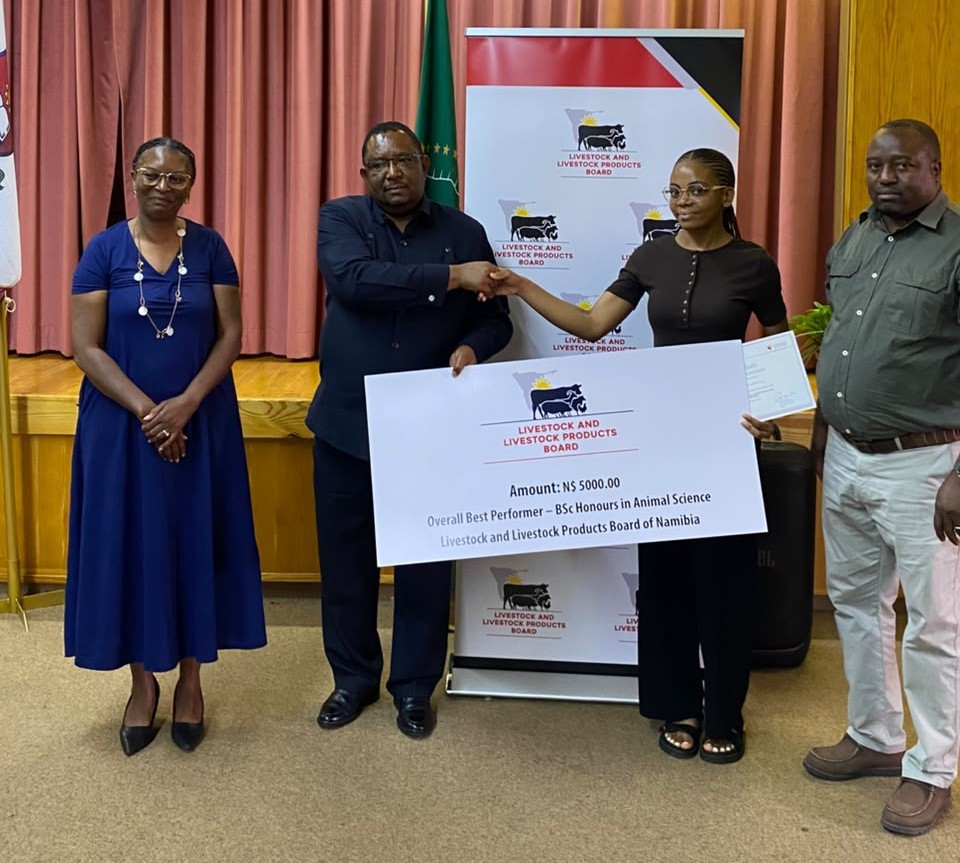 Award ceremony scene: two men shake hands as a woman presents a large check from the Livestock and Livestock Products Board of Namibia, with a banner behind them.