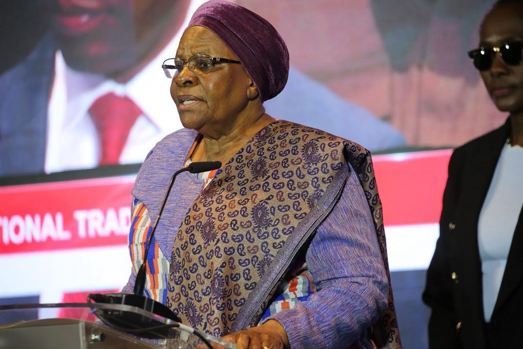 PRESIDENT4 - The Brief | Namibia's Leading Business & Financial News Elderly African woman in a purple headscarf and patterned outfit speaking at a podium with a microphone on stage.