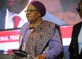 Elderly African woman in a purple headscarf and patterned outfit speaking at a podium with a microphone on stage.