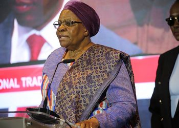 PRESIDENT4 - The Brief | Namibia's Leading Business & Financial News Elderly African woman in a purple headscarf and patterned outfit speaking at a podium with a microphone on stage.