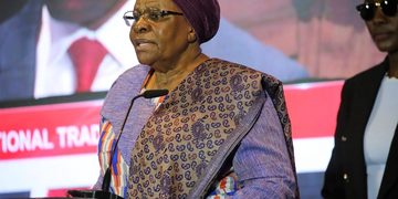 Elderly African woman in a purple headscarf and patterned outfit speaking at a podium with a microphone on stage.