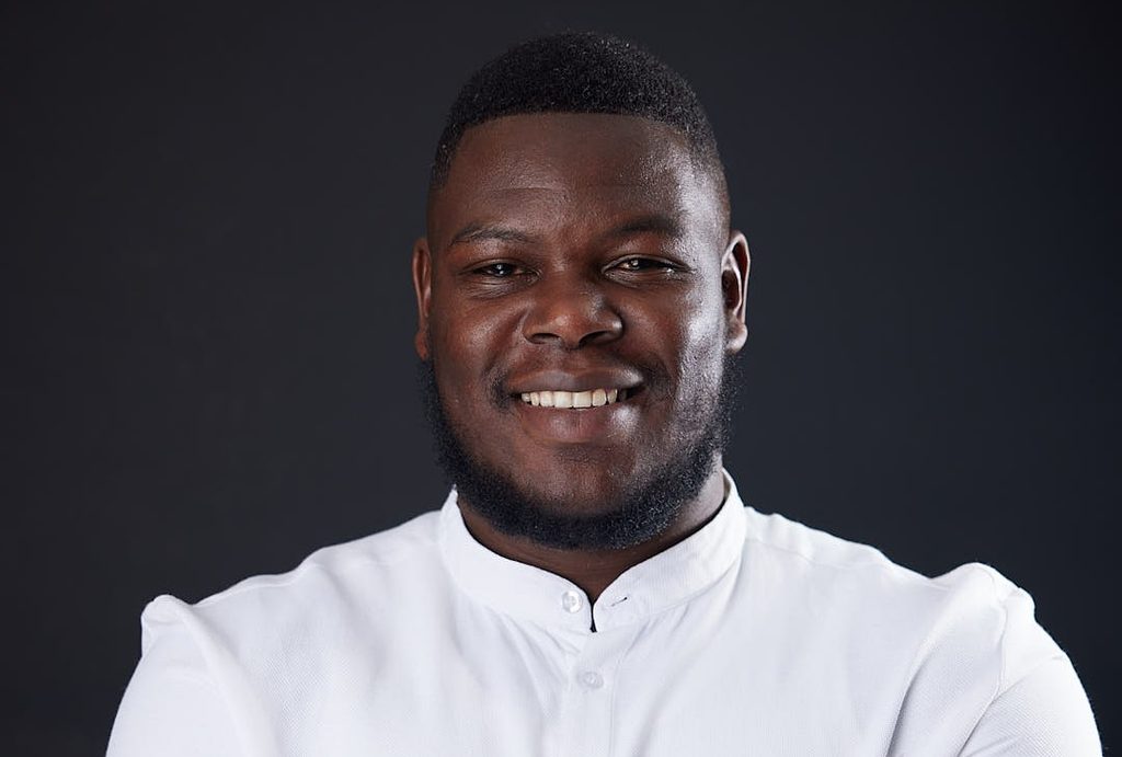 Close-up portrait of a smiling Black man in a white button-up shirt against a dark backdrop.