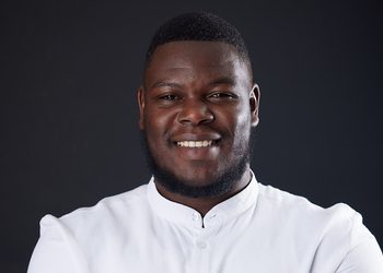 Close-up portrait of a smiling Black man in a white button-up shirt against a dark backdrop.
