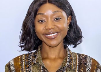 Portrait of a smiling woman with shoulder-length dark hair wearing a brown and green patterned blouse against a light background.
