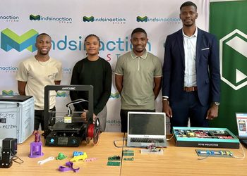Group of four people standing behind a table with 3D printers and electronics at a tech exhibit, branded banners in the background.