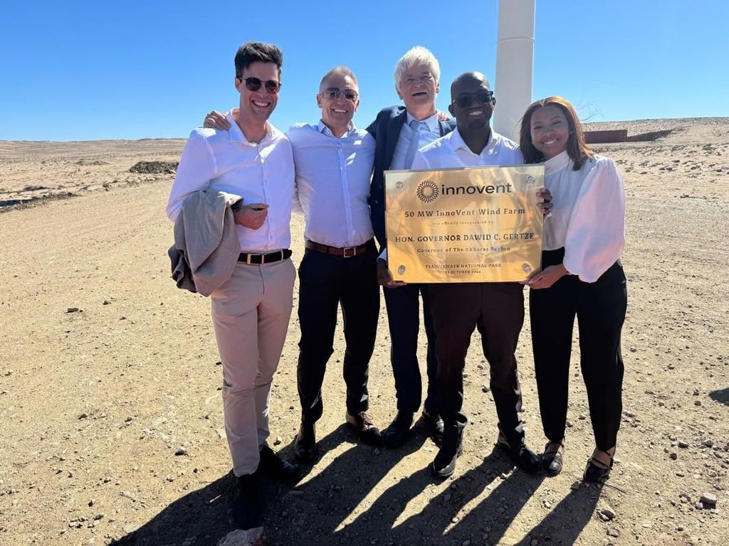 Five adults in smart attire posing outdoors with a plaque at the Innovent Wind Farm site in a desert under a clear blue sky.