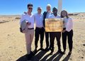 Five adults in smart attire posing outdoors with a plaque at the Innovent Wind Farm site in a desert under a clear blue sky.