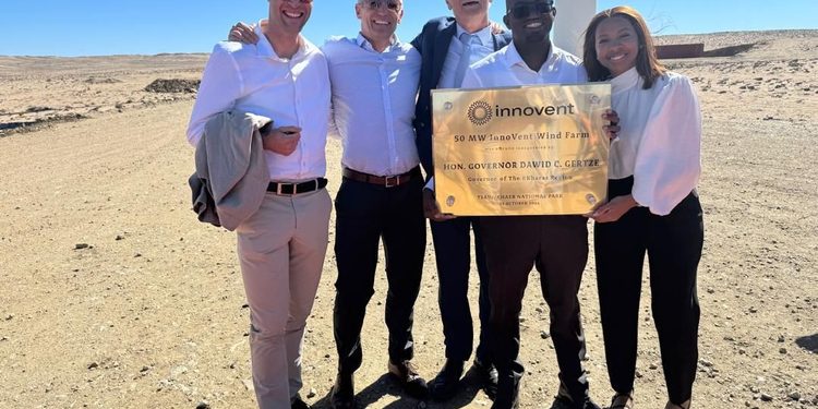 Five adults in smart attire posing outdoors with a plaque at the Innovent Wind Farm site in a desert under a clear blue sky.