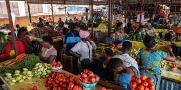 Bustling open-air market with stalls of tomatoes, peppers, and greens as shoppers browse and interact under a corrugated roof.