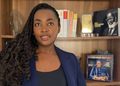 Woman with long curly hair in a navy blazer stands in front of a wooden bookshelf lined with books and photos.