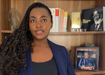 Woman with long curly hair in a navy blazer stands in front of a wooden bookshelf lined with books and photos.