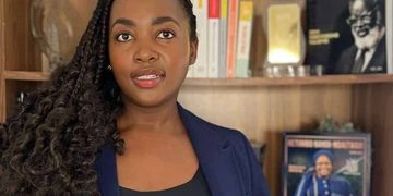 Woman with long curly hair in a navy blazer stands in front of a wooden bookshelf lined with books and photos.