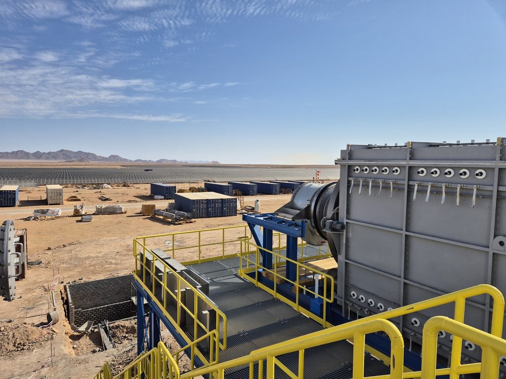 Industrial facility in a desert with large metal equipment, yellow safety railings, and rows of solar panels in the distance under a clear blue sky.