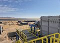 Industrial facility in a desert with large metal equipment, yellow safety railings, and rows of solar panels in the distance under a clear blue sky.