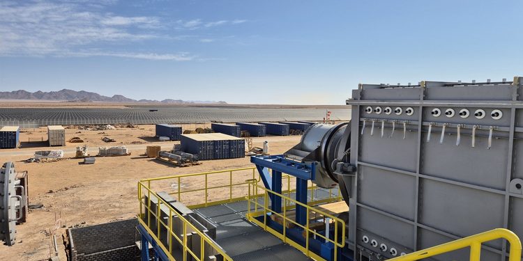 Industrial facility in a desert with large metal equipment, yellow safety railings, and rows of solar panels in the distance under a clear blue sky.