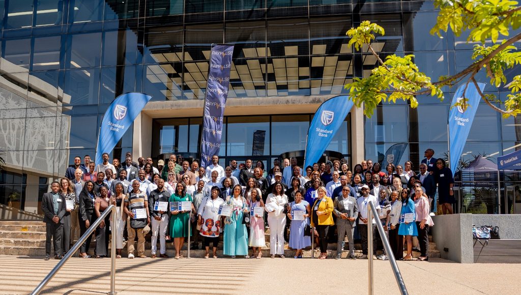 WhatsApp Image 2026-10-29 at 104707 AM - The Brief | Namibia's Leading Business & Financial News Large diverse group of professionals on building steps, certificates in hand, at an event outside a glass-fronted venue with Standard Bank banners.