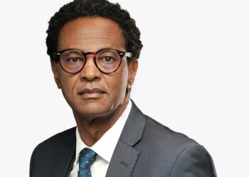 Professional man in a dark gray suit and blue tie, wearing round glasses, looking straight at the camera.