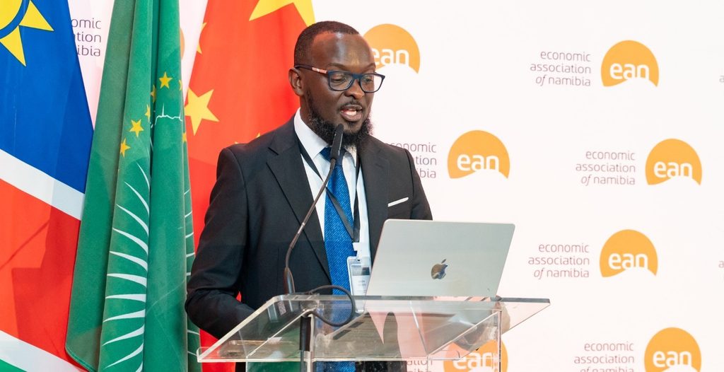 JASON KASUTO1 - The Brief | Namibia's Leading Business & Financial News A man in a dark suit and blue tie speaks at a clear podium with a laptop, microphone, and event backdrop displaying 'economic association of Namibia' logos and flags behind him.