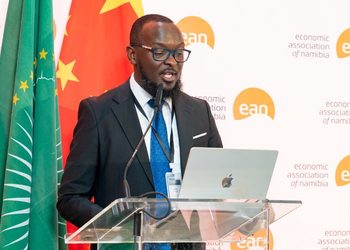 A man in a dark suit and blue tie speaks at a clear podium with a laptop, microphone, and event backdrop displaying 'economic association of Namibia' logos and flags behind him.