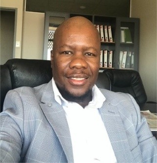 Smiling man in a light gray checkered blazer and white shirt, seated in an office with shelves and binders behind him.