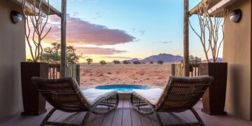 Two wicker lounge chairs on a wooden deck overlook a small circular pool in a desert landscape at sunset.
